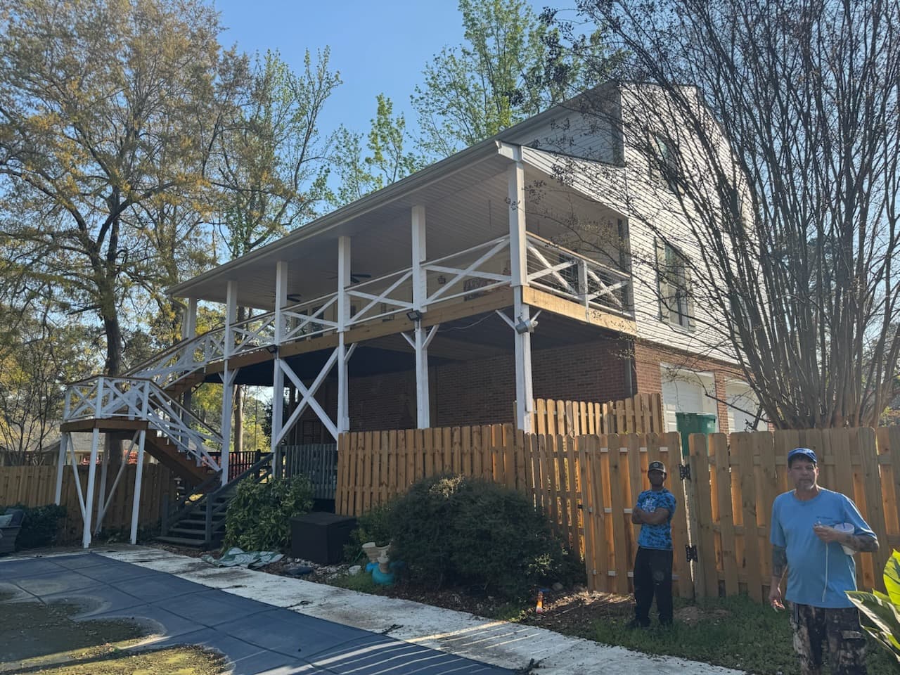 White elevated deck with cross railings attached to a brick house behind a wooden fence.